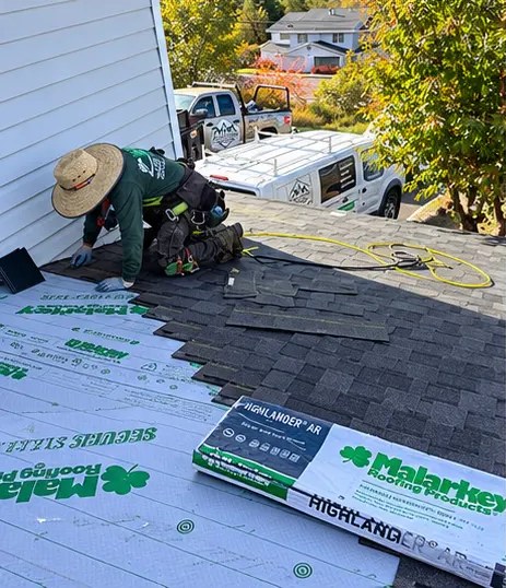 Everguard roofer in a sun hat installing dark Malarkey shingles beside bundles of Malarkey Highlander shingles