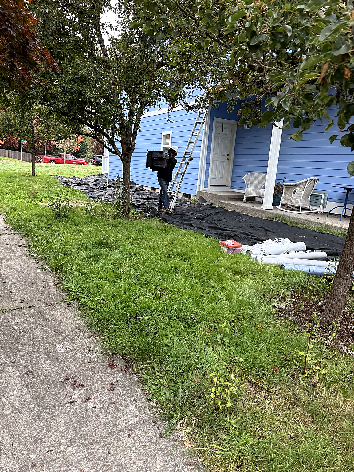 Roofing crew member carrying removed roofing debris across protective tarps beside a blue house and ladder