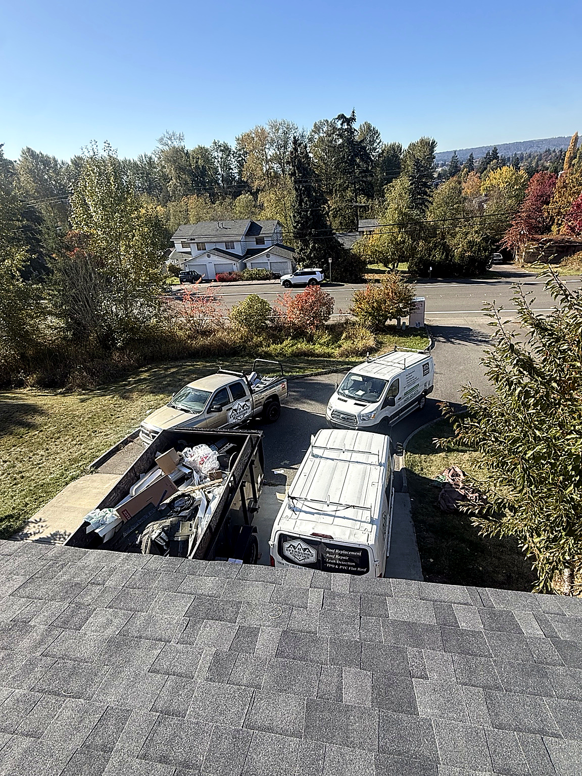 View from the roof over new shingles toward Everguard work vans, pickup and debris trailer in the driveway
